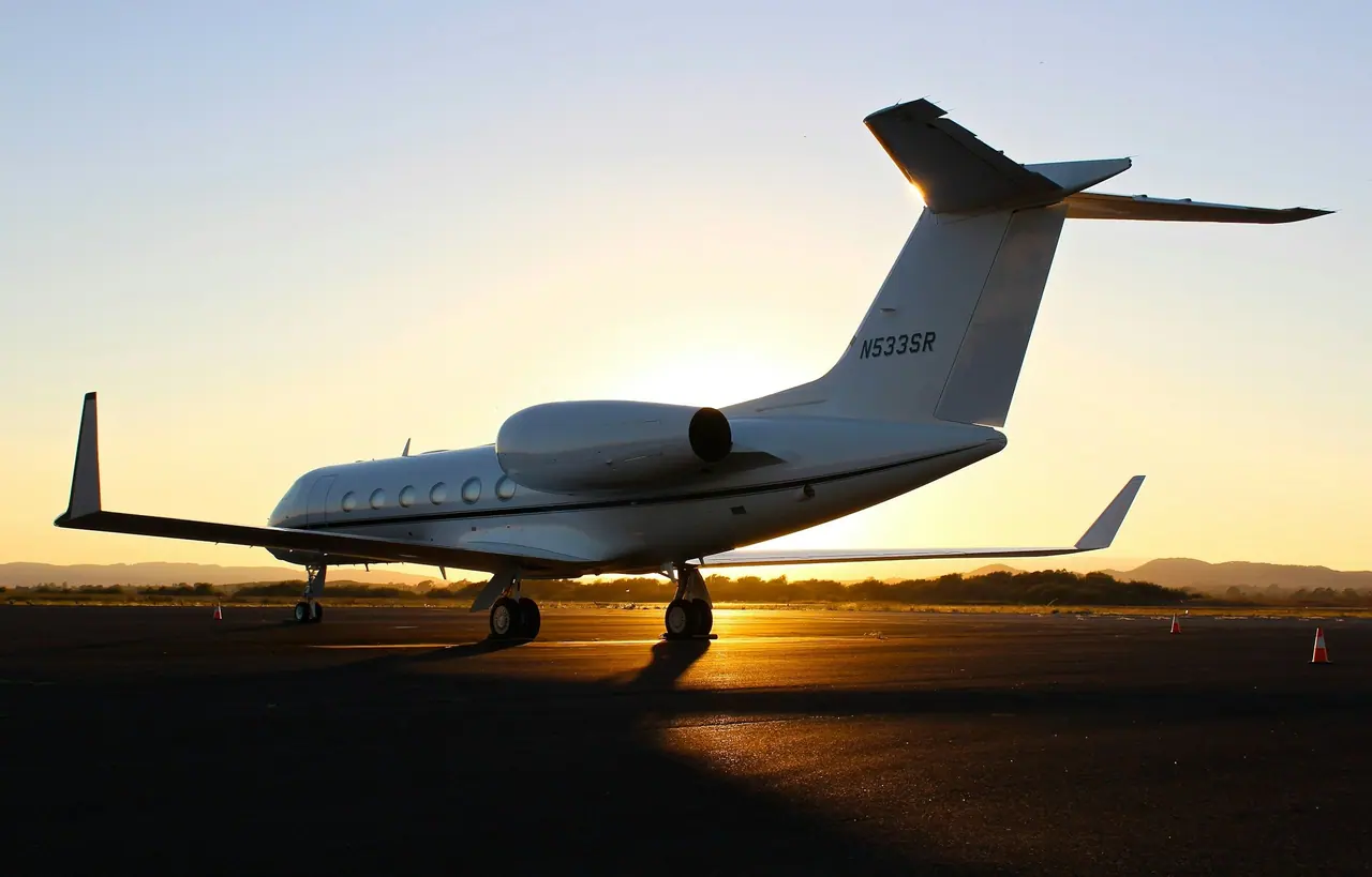 A white business jet parked on the ramp at sunset, low sun glowing beneath its wing